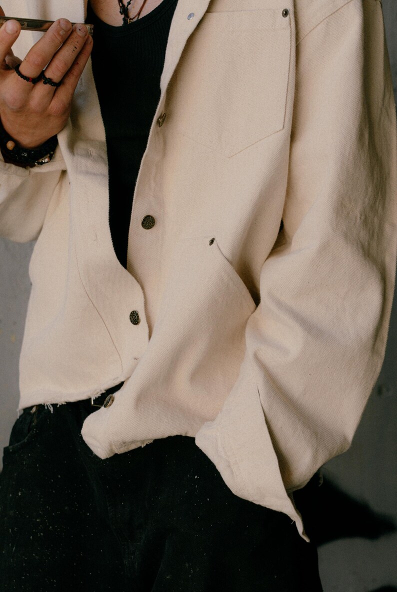 May include: A long-sleeved, off-white button-down shirt made of a heavy fabric, featuring a chest pocket, a pointed collar, and metallic buttons. The shirt has a raw, unfinished hemline at the bottom and is worn over a plain black undershirt.