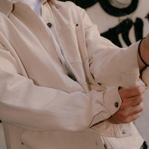 May include: An off-white long-sleeve button-down shirt featuring a classic collar, a single chest pocket, and metallic buttons. The shirt is worn over a plain white t-shirt, showcasing a structured fabric with visible stitching details on the cuffs.