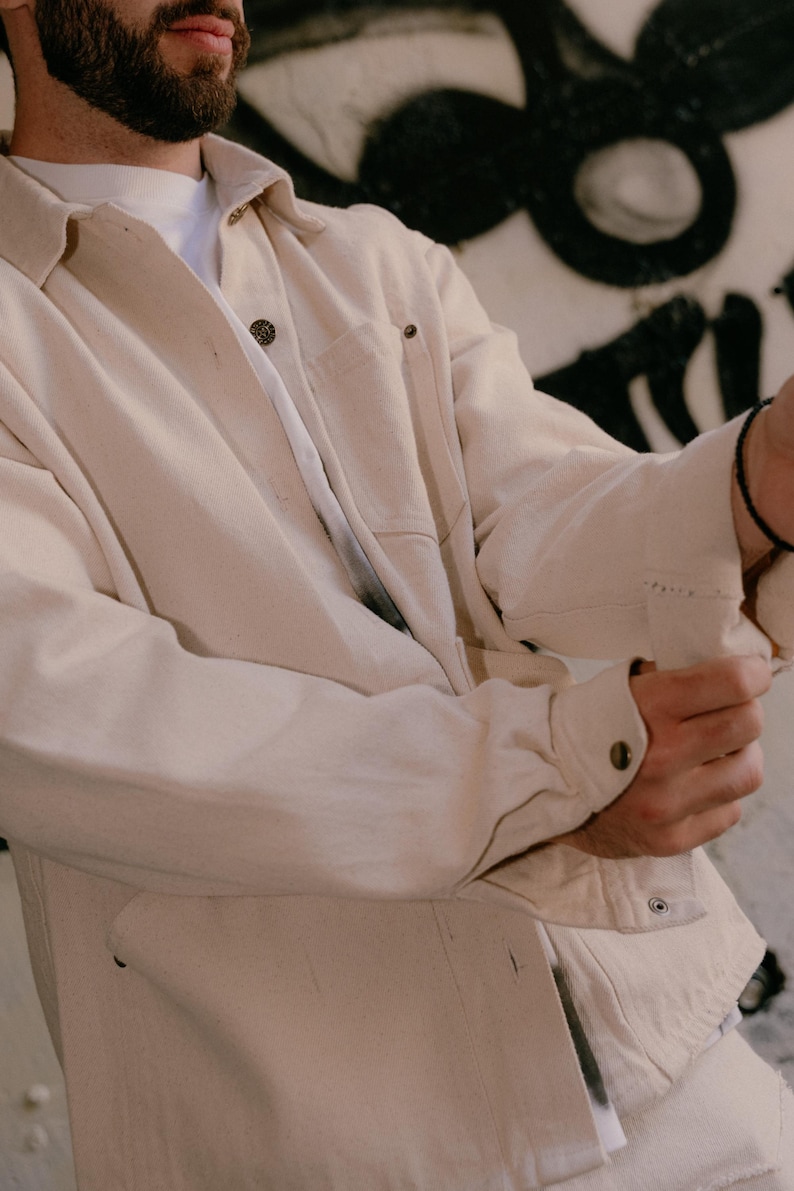 May include: An off-white long-sleeve button-down shirt featuring a classic collar, a single chest pocket, and metallic buttons. The shirt is worn over a plain white t-shirt, showcasing a structured fabric with visible stitching details on the cuffs.