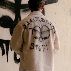 May include: A beige long-sleeve button-up shirt with a frayed circular patch on the back featuring a black spray-painted design. The text "BLUEBERRY STUFF" is spray-painted in black around the patch. Matching beige trousers feature black graffiti.