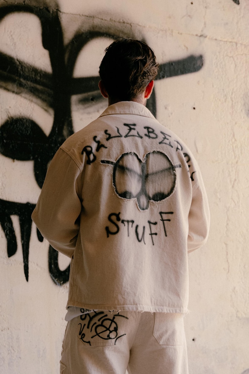 May include: A beige long-sleeve button-up shirt with a frayed circular patch on the back featuring a black spray-painted design. The text "BLUEBERRY STUFF" is spray-painted in black around the patch. Matching beige trousers feature black graffiti.