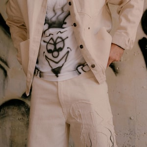 May include: A beige button-down jacket worn over a white t-shirt with a black airbrushed cartoon face graphic. The outfit includes matching beige trousers featuring a large, frayed-edge fabric patch on the left leg with black airbrushed details.