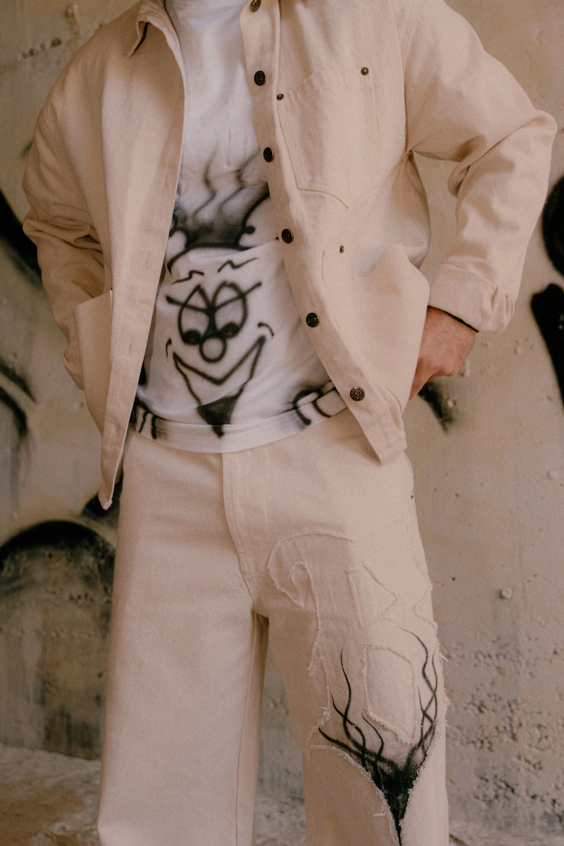 May include: A beige button-down jacket worn over a white t-shirt with a black airbrushed cartoon face graphic. The outfit includes matching beige trousers featuring a large, frayed-edge fabric patch on the left leg with black airbrushed details.