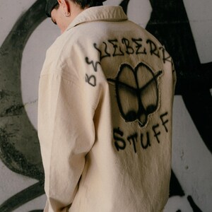May include: A beige long-sleeve button-down shirt featuring black spray-painted graffiti on the back. The design includes the text "ICEBERG STUFF" surrounding a central abstract shape with frayed edges. The shirt has a collar and long sleeves.