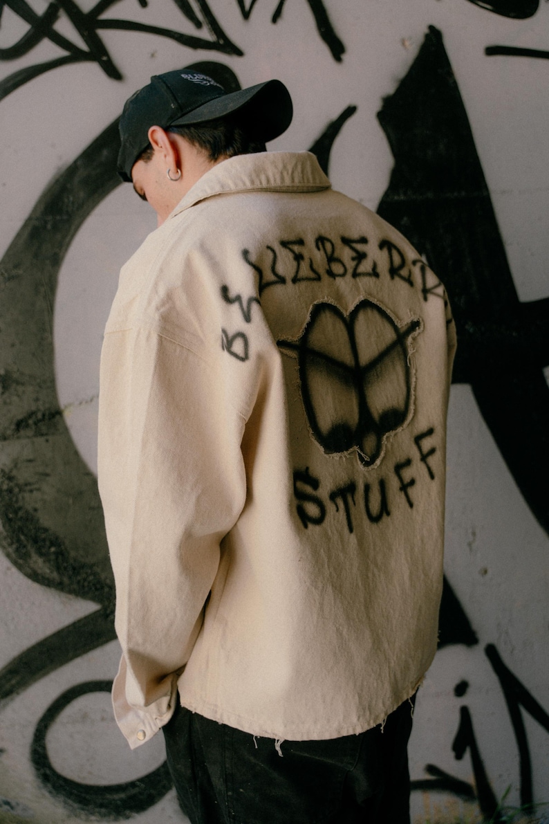 May include: A beige long-sleeve button-down shirt featuring black spray-painted graffiti on the back. The design includes the text "ICEBERG STUFF" surrounding a central abstract shape with frayed edges. The shirt has a collar and long sleeves.