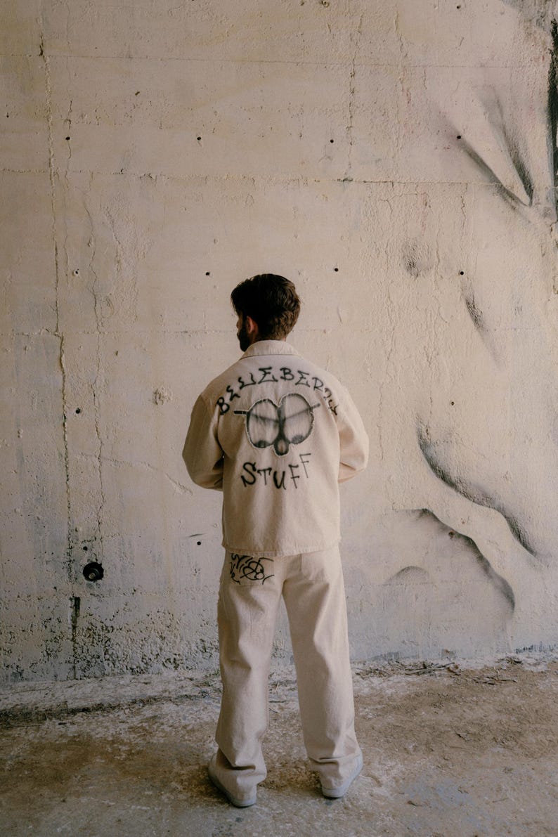 May include: A beige long-sleeved button-up shirt and matching trousers. The back of the shirt features a black graphic of two eyes and the text "BLUEBERRY STUFF" in a handwritten style. The trousers have a small black graphic on the back pocket.