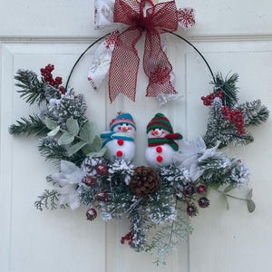 Puede incluir: Una festiva corona navideña con dos muñecos de nieve con sombreros y bufandas coloridas. La corona está decorada con ramas de pino artificiales, bayas rojas y una piña. Un lazo rojo y blanco con detalles de copos de nieve remata la corona.