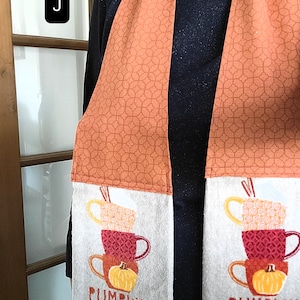 May include: A kitchen towel with a fall-themed design. It features a stack of mugs with a pumpkin on top and the text "Pumpkin Spice and everything nice". The towel is orange and white with a geometric pattern.