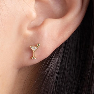 Puede incluir: Pendientes de vidrio de martini en tono dorado con un pequeño acento de piedra preciosa verde. Los pendientes son pequeños y delicados, perfectos para el uso diario.