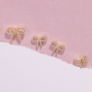 Puede incluir: Seis pendientes dorados en forma de lazo con piedras transparentes y brillantes están dispuestos sobre un fondo rosa. Los pendientes varían ligeramente de tamaño, con tres pares visibles. Los lazos son detallados y delicados.