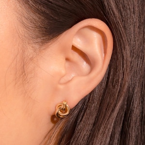 May include: Close-up of a gold-colored knot stud earring. The earring is a small, round design with a textured surface. The earring is worn on the ear of a person with brown hair.