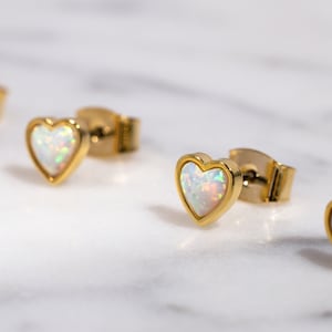 May include: A set of four gold heart-shaped earrings with white opal stones. The earrings are on a white marble surface.