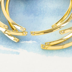 Op de afbeelding: Goudkleurige sieraden met een gebogen ontwerp. Elk stuk heeft kleine, vierkant geslepen edelstenen. De sieraden zijn geplaatst tegen een lichtblauwe en witte achtergrond, wat een aantrekkelijk contrast creëert.