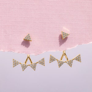 May include: Gold-toned earrings with a geometric design. The earrings feature a triangle stud and a larger, decorative piece with multiple triangle shapes, all encrusted with small, sparkling stones. The earrings are displayed on a pink textured background.