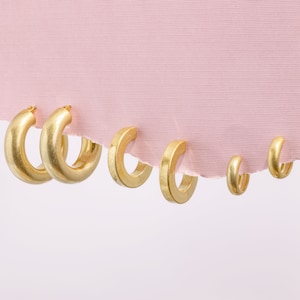 May include: Five gold hoop earrings of varying sizes, hanging from a pink fabric background.