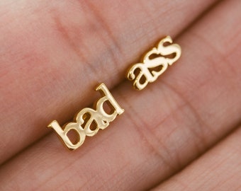 May include: Gold-colored stud earrings spelling out "bad ass" in a cursive font. The earrings are small and delicate, with a polished finish. The letters are slightly rounded and connected.