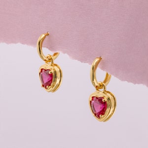 May include: A pair of gold hoop earrings with a dangling heart-shaped charm. Each charm features a red gemstone.