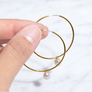 Puede incluir: Pendientes de aro dorados con una pequeña perla colgante. Los aros están sostenidos por una mano sobre un fondo de mármol blanco. Los pendientes tienen un diseño simple y elegante.