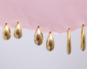 May include: Five gold-colored earrings with different shapes and sizes. The earrings are arranged in a row on a white and pink surface.