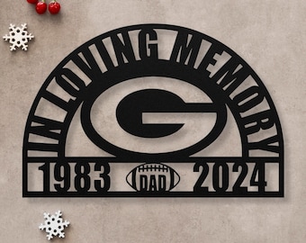 May include: Black metal memorial sign featuring a green and white Green Bay Packers logo, the words "In Loving Memory", the years "1983" and "2024", and a football with the word "Dad" on it.