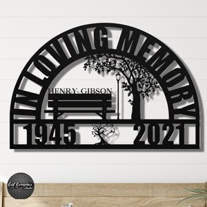 May include: Black metal memorial sign with the words "In Loving Memory" in a half-circle arch. The sign features a silhouette of a bench, a tree, and a lamppost. The name "HENRY GIBSON" is inscribed on the bench, and the dates "1945" and "2021" are displayed below the bench.