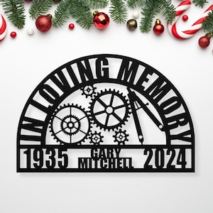May include: Black metal memorial sign with gears and a compass design. The sign reads "In Loving Memory" with the years "1935" and "2024" and the name "Gary Mitchell" in the center.