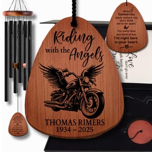 May include: A wooden wind chime with black chimes and a decorative wooden plaque. The plaque features a motorcycle with angel wings and the text "Riding with the Angels" and a name and dates. A smaller plaque with a poem hangs nearby.