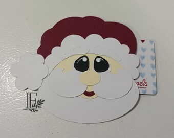 May include: A white paper craft of Santa Claus's face with a red hat, white beard, and black eyes. The craft is on a white background with a small tag that says "Michaels".