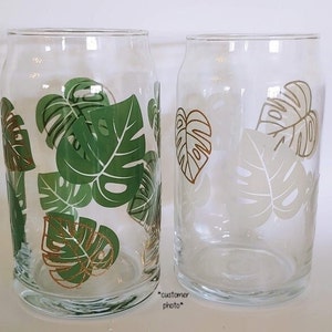 May include: Two clear glass drinking glasses with a tropical leaf design. The glasses have green and gold leaves with a white outline.
