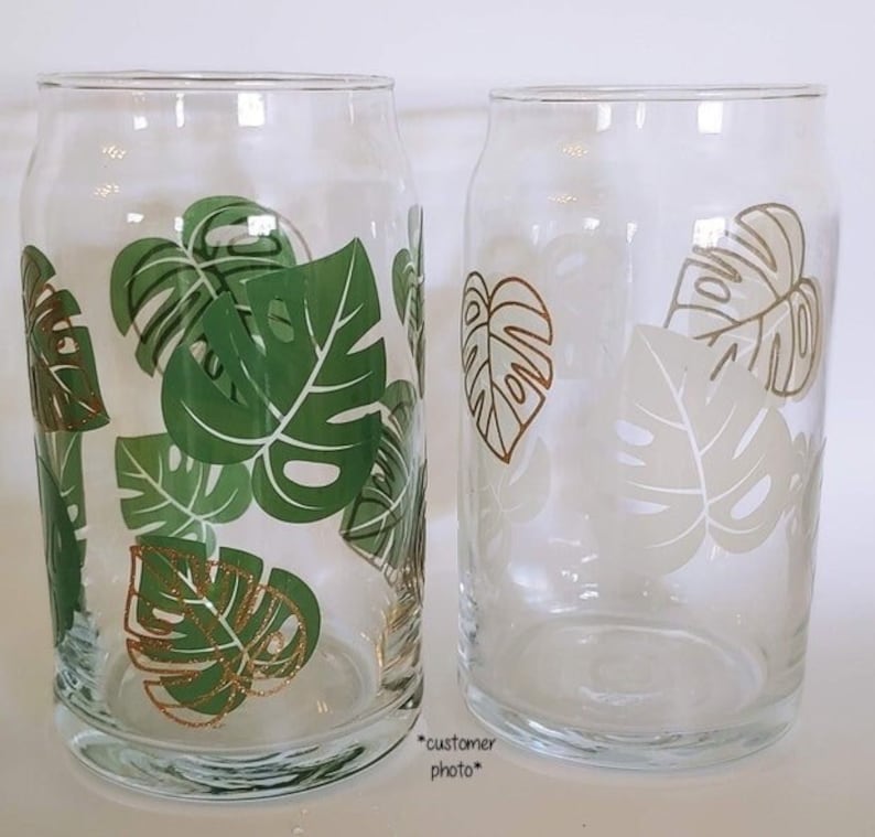 May include: Two clear glass drinking glasses with a tropical leaf design. The glasses have green and gold leaves with a white outline.