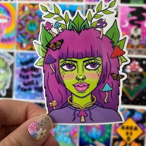 May include: A colorful sticker featuring a stylized illustration of a woman with green skin, purple hair, and mushroom and butterfly accents. The woman is wearing a purple top and a gold necklace. The background is filled with other colorful stickers.