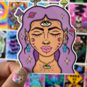 May include: A sticker featuring a purple-haired woman with a third eye, surrounded by mushrooms and stars. The woman has a gold necklace with an eye pendant and is wearing earrings with blue crystals.