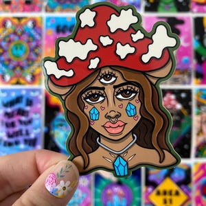 May include: A sticker featuring a cartoon illustration of a person with a third eye, wearing a red and white mushroom cap. The person has blue teardrop-shaped crystals on their cheeks and a blue crystal pendant around their neck.