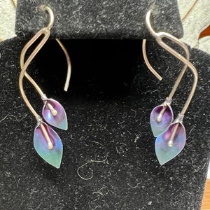 May include: A pair of silver earrings with a delicate, twisted design. Each earring features two iridescent purple and green flower-shaped charms.