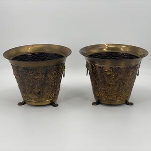 Peut inclure: Deux jardinières antiques en laiton avec des motifs floraux ornés. Chaque jardinière a un large bord, de petites poignées et trois petits pieds. L'intérieur est sombre et l'extérieur a une patine chaude et vieillie.