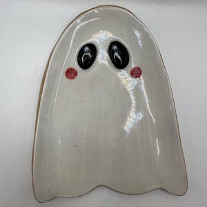 May include: A ghost-shaped ceramic tray with a glossy off-white finish and speckled texture. The tray features two raised, smooth black oval eyes and two small, circular red painted cheeks on the face, with a wavy bottom edge.