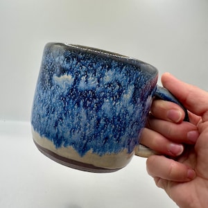 Peut inclure: Une tasse en céramique avec un émail réactif présentant un motif moucheté bleu marine profond et blanc qui se transforme en une base beige unie. La tasse possède une anse épaisse et arrondie ainsi qu'un bord sombre non émaillé.