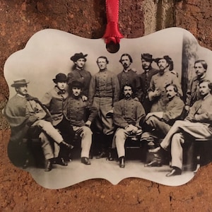 May include: Ornament featuring a black and white photograph of a group of men in military uniforms. The photograph is in an ornate, irregular shape, with a red ribbon for hanging. The men are posed in a group, some seated and some standing.