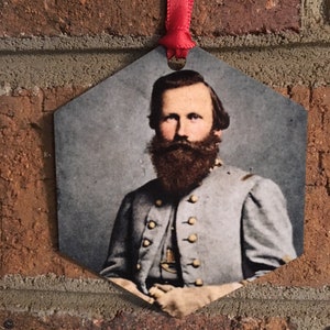 May include: A hexagonal ornament featuring a portrait of a man with a long beard and gray uniform. The image is attached to a red ribbon for hanging. The background is a brick wall.