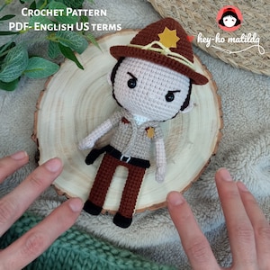 May include: Crochet pattern for a brown and beige amigurumi doll wearing a brown hat with a yellow star. The doll has a stern expression and is holding a brown gun. The doll is sitting on a wooden surface. The text "hey-ho matilda" is written in red and white on the background.