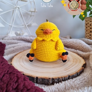 Könnte beinhalten: Ein handgehäkeltes gelbes Küken mit orangefarbenen und schwarzen Füßen und einem kleinen Schnabel. Das Küken sitzt auf einer Holzscheibe. Das Bild enthält den Text "Tutie Tutie" und ein Logo mit Würfel und Häkelhaken.