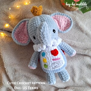 以下が含まれることがあります： 王冠をかぶった、かぎ針編みで作ったぬいぐるみのおもちゃの象。白と青のベストを着て、赤いハートとカラフルなドットが描かれています。おもちゃは薄い茶色のふわふわした表面の上に置かれています。