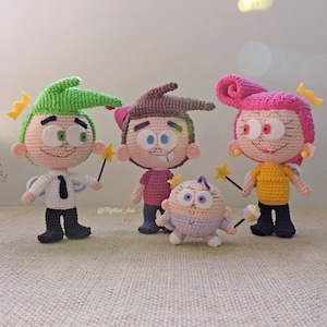 Puede incluir: Figuras de ganchillo de personajes de dibujos animados, incluyendo una figura de pelo verde con camisa blanca, una figura de pelo castaño con camisa rosa, una figura de pelo rosa con camisa amarilla y una pequeña figura morada. Cada figura sostiene una varita.