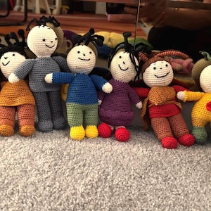 Pode incluir: Uma coleção de dez bonecos de crochê feitos à mão com várias roupas coloridas, incluindo peças vermelhas, roxas, amarelas, azuis e marrons. Cada boneco tem uma cabeça redonda branca com traços faciais bordados e cabelo de lã preta.