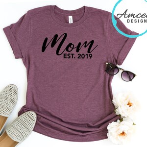 May include: A maroon t-shirt with the text "Mom Est. 2019" in black lettering.