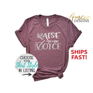 May include: A maroon t-shirt with the text "RAISE YOUR VOICE" in silver glitter lettering. The shirt has a rolled cuff sleeve and is made of a soft, comfortable fabric.