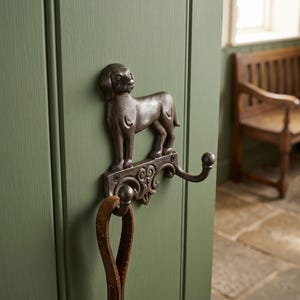 May include: A dark brown cast iron wall hook shaped like a standing dog. The hook features a decorative scrollwork base with two mounting screws and two curved prongs for hanging items. A brown leather leash hangs from one of the hooks.