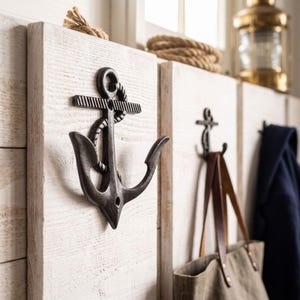 May include: A wall-mounted coat rack featuring white-washed wooden planks and dark metal anchor-shaped hooks. The hooks are designed with a rope detail wrapped around the anchor shaft. A canvas bag with leather straps hangs from one of the hooks.