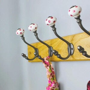 May include: A wall-mounted coat rack featuring a light-coloured wooden board with four dark metal hooks. Each hook is topped with a white ceramic knob decorated with small pink polka dots. A patterned pink and green scarf hangs from one hook.