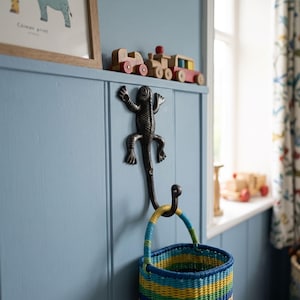 May include: A dark brown, cast iron wall hook shaped like a lizard with a curved tail that functions as a hook. A woven basket with horizontal stripes of blue, yellow, and green is hanging from the tail of the lizard-shaped wall hook.